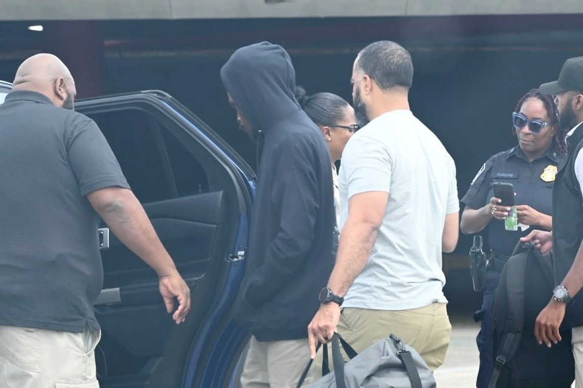 Jahi Grant, wearing a dark hoodie, is escorted toward a vehicle by law enforcement personnel. Grant was returned to St. Thomas after his arrest in Dominica to face 12 firearm, drug and assault-related charges.